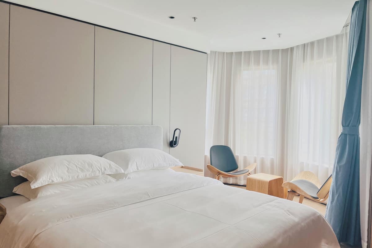A modern hotel room with a large bed, a seating area with chairs, and a view of the outdoors through sheer curtains.