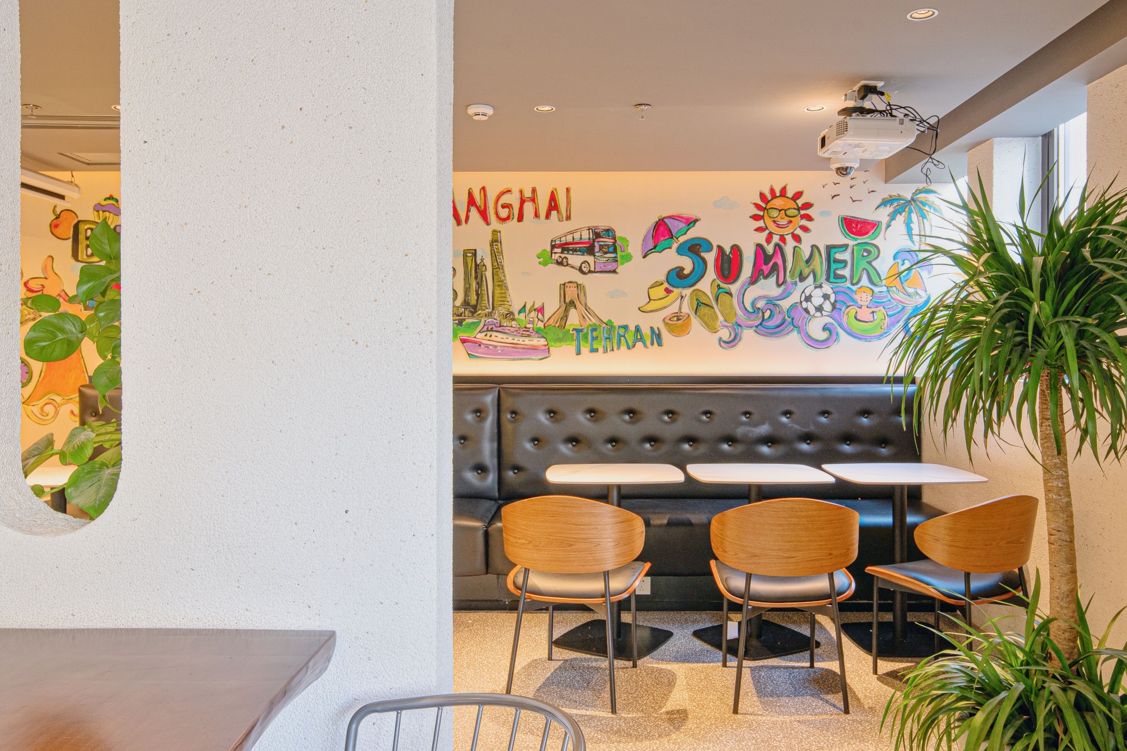 A restaurant with a wall mural, a black cushioned booth, wooden chairs, and a potted plant.