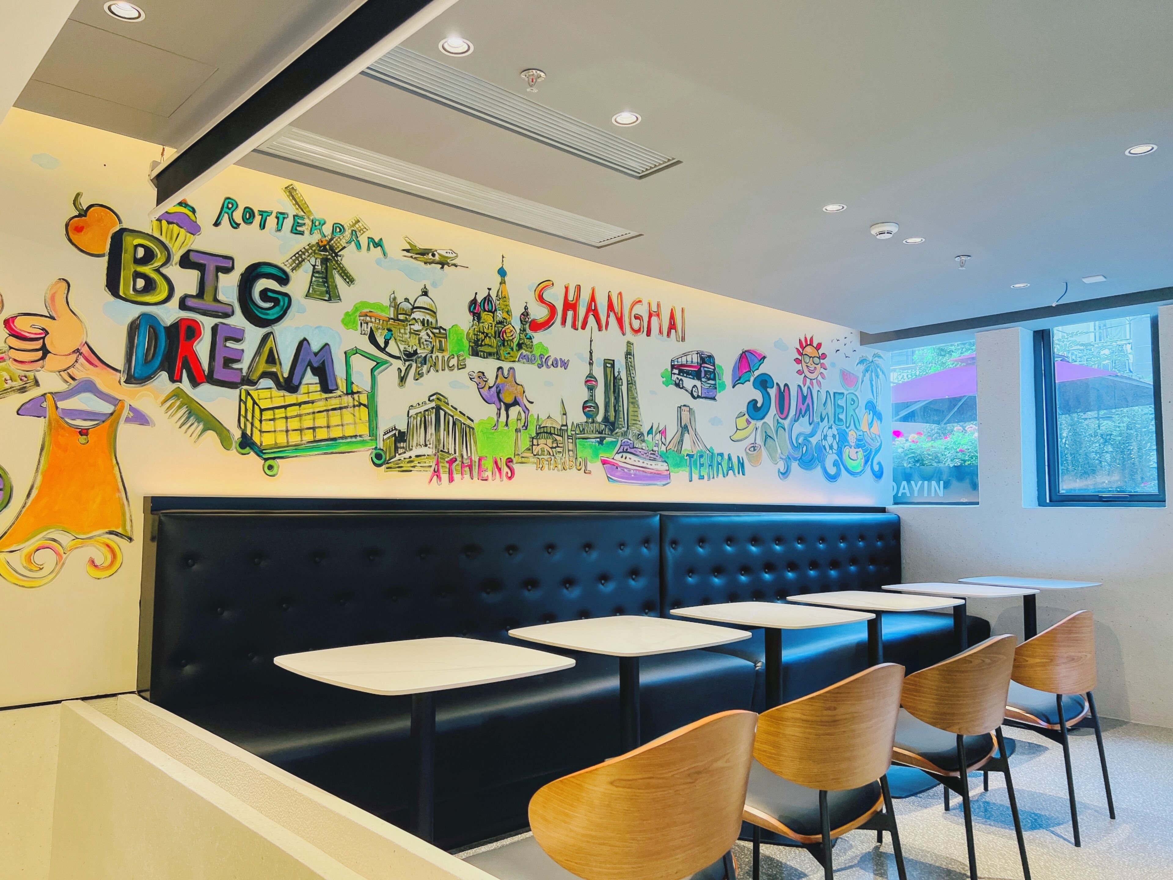 A restaurant interior with a wall mural featuring travel destinations, a black cushioned booth seating area, and wooden chairs.