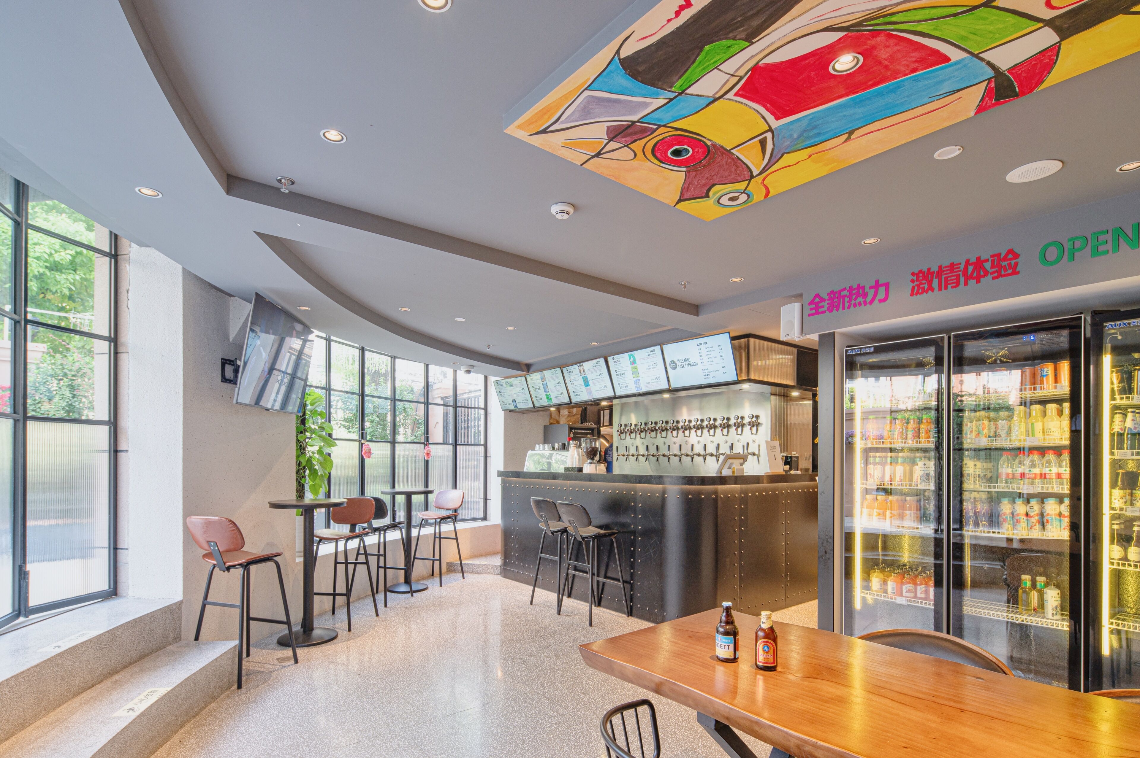 A modern bar area with seating, a refrigerated display case, and a colorful ceiling artwork.