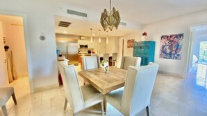 A modern dining area with a wooden table and white chairs, a kitchen in the background, and a blue cabinet with artwork on the wall.