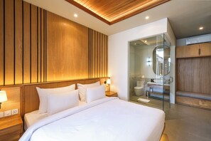 A modern hotel room with a large bed, wooden panel wall, and a glass-enclosed bathroom. - Villa Aashiyana (Canggu)