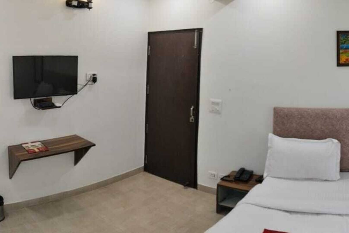 Hotellihuone, jossa on sänky, seinälle asennettu televisio, sängyn yläpuolella oleva taulu, ovi sekä yöpöytä, jossa on puhelin ja kahvinkeitin.