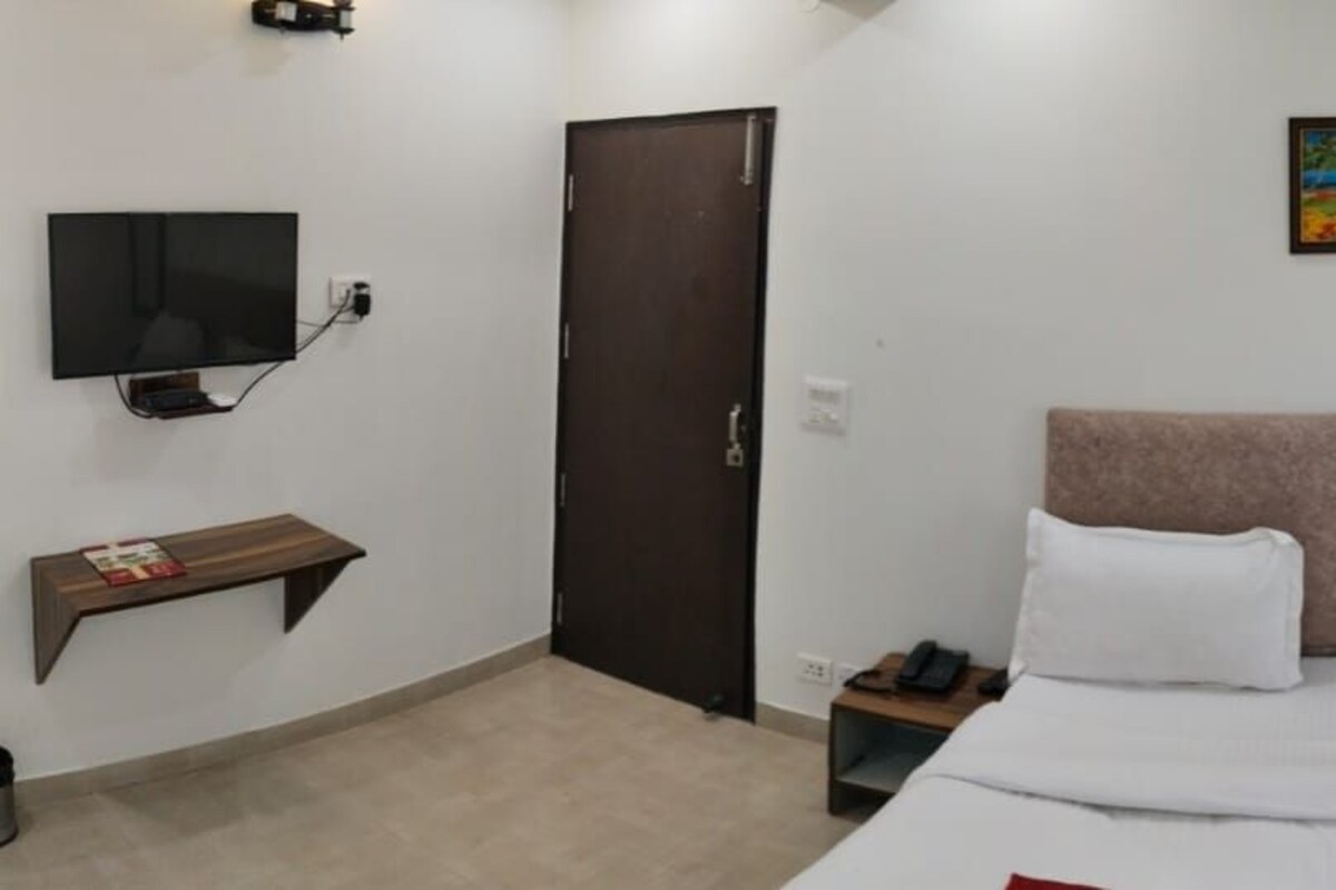 Une chambre d’hôtel avec un lit, un téléviseur fixé au mur, un tableau au-dessus du lit, une porte et une table de chevet avec un téléphone et une machine à café.
