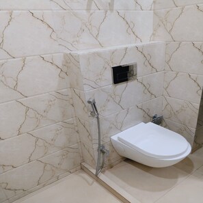 A bathroom with a white toilet, a shower with a handheld showerhead, and a marble-patterned wall. - Fortune Residency (New Delhi)