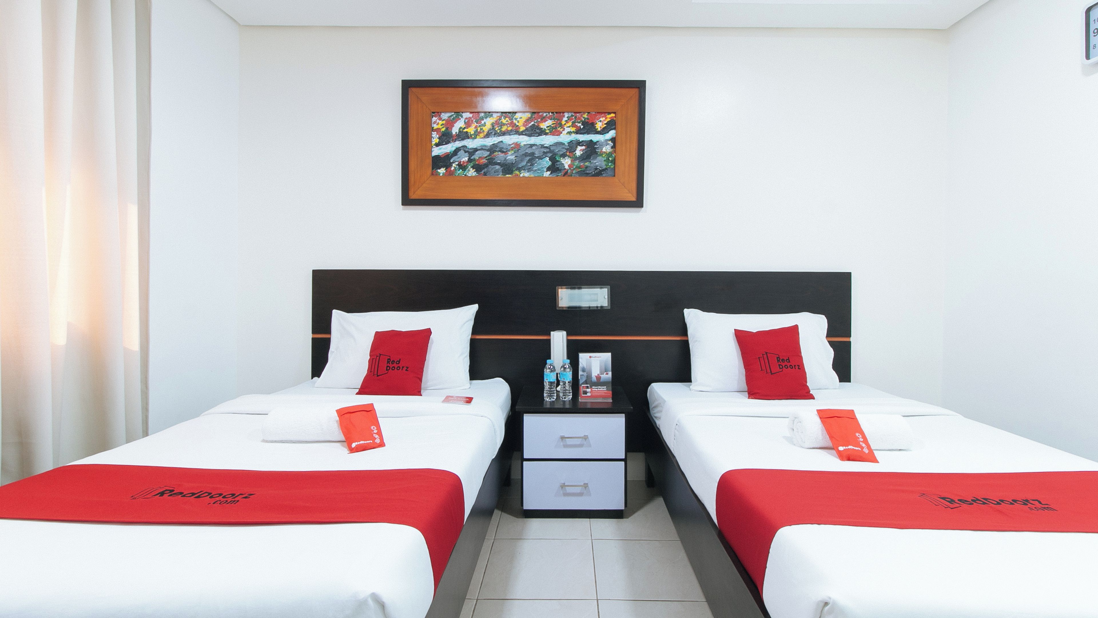 A hotel room with two beds, each with white bedding and red accents, a framed artwork above the beds, and a small table between the beds.