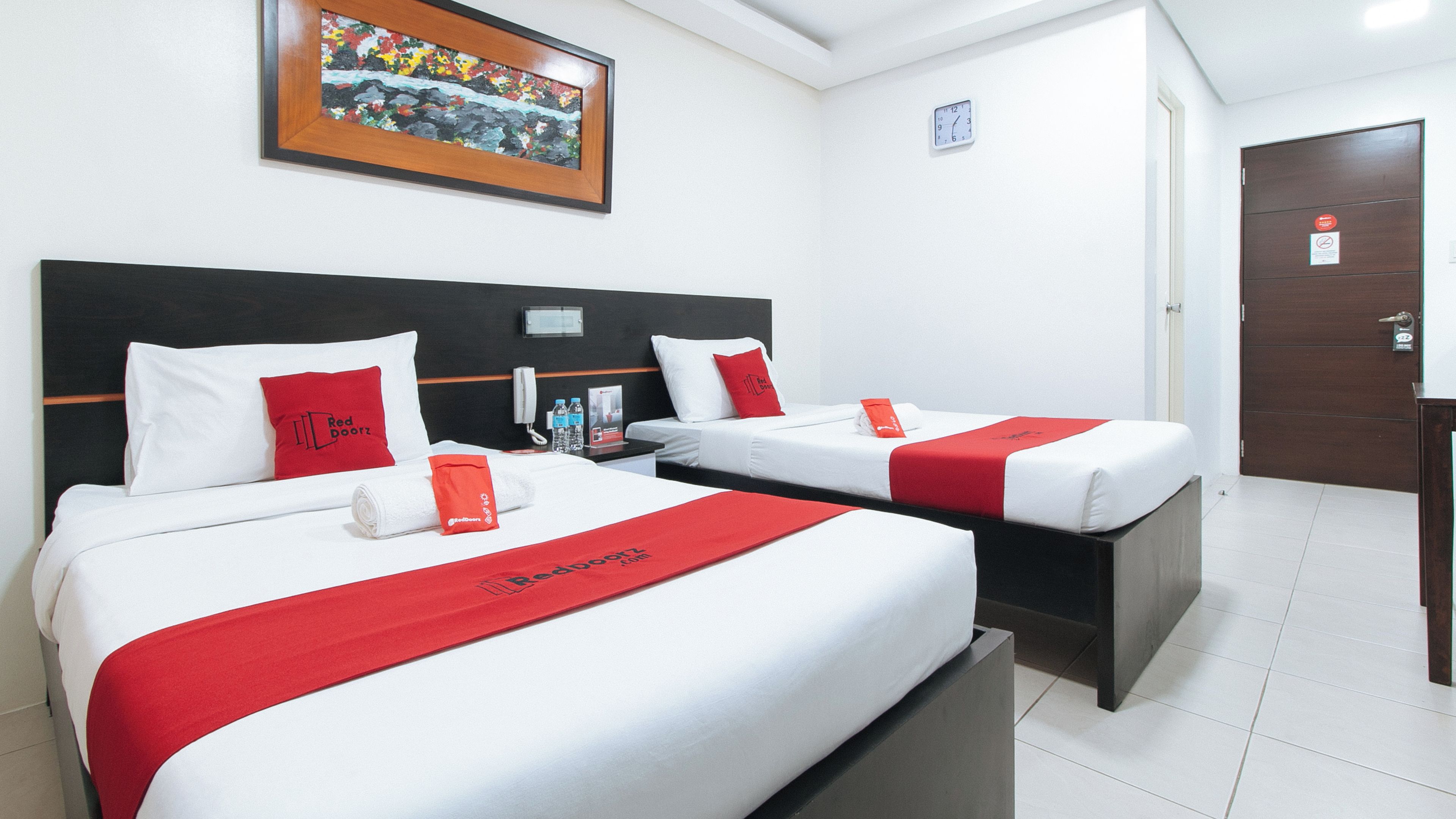 A hotel room with two beds, each with white bedding and red accents, a painting above the beds, a clock on the wall, and a door with a sign.