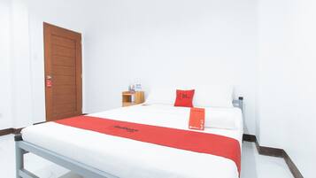 A hotel room with a bed, a wooden door, a small table with a red pillow, and a white wall.