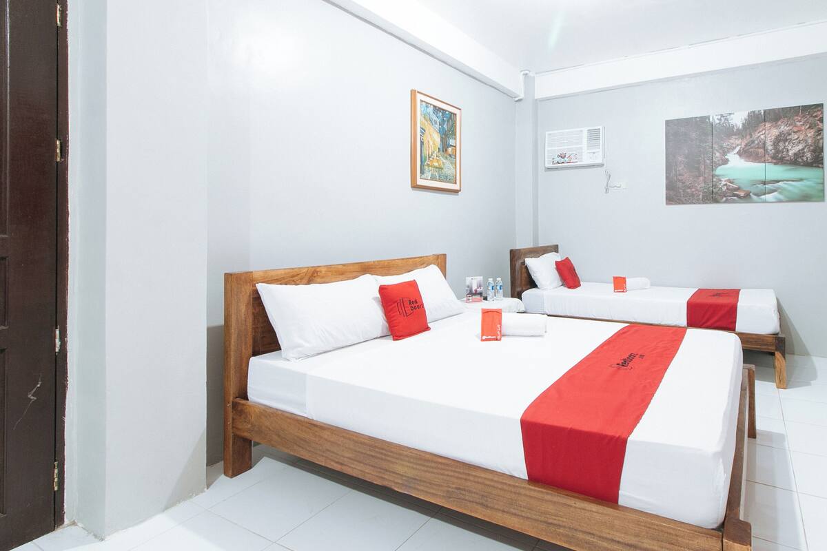 Ein Hotelzimmer mit zwei Betten, weißer Bettwäsche, roten Akzenten, einem Gemälde an der Wand und einem Fenster mit Klimaanlage.