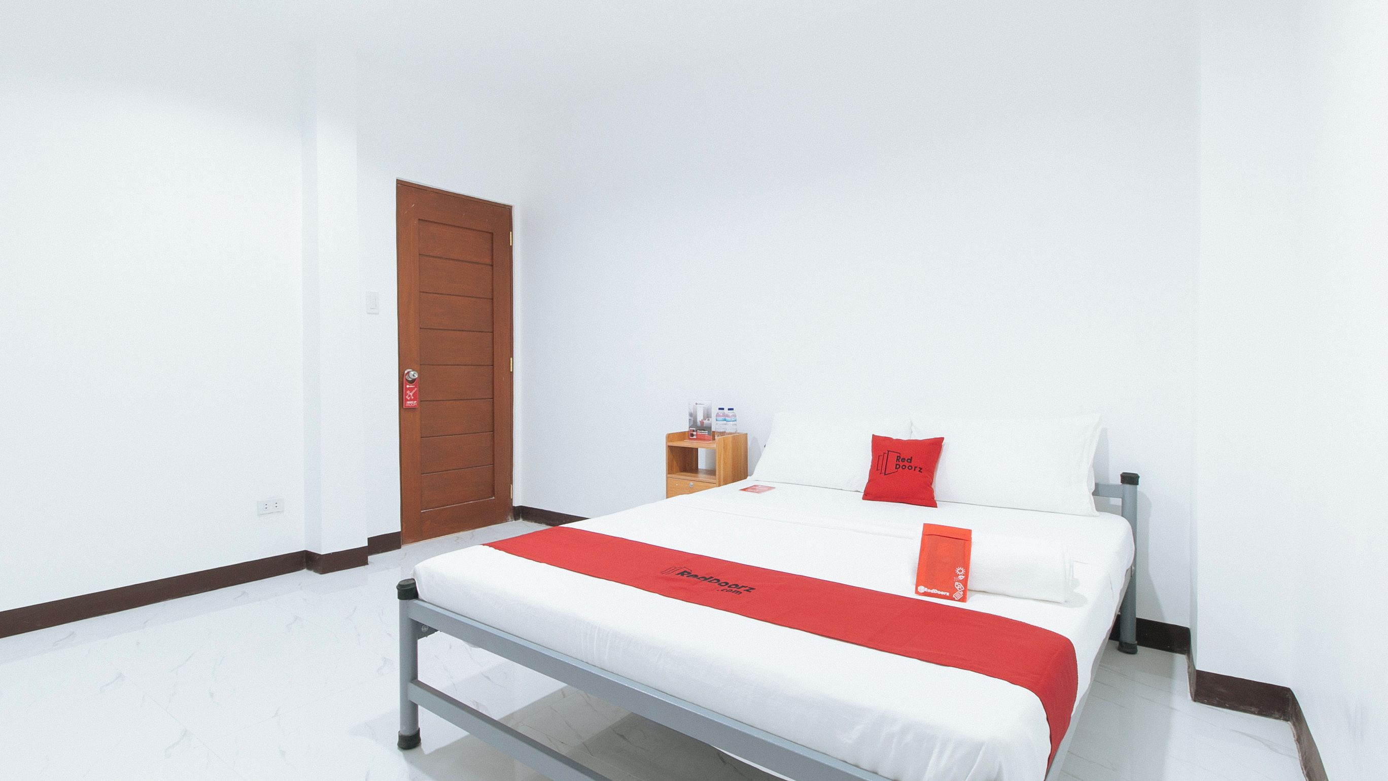 A hotel room with a bed, a wooden door, a small wooden nightstand, and a red and white bedspread.