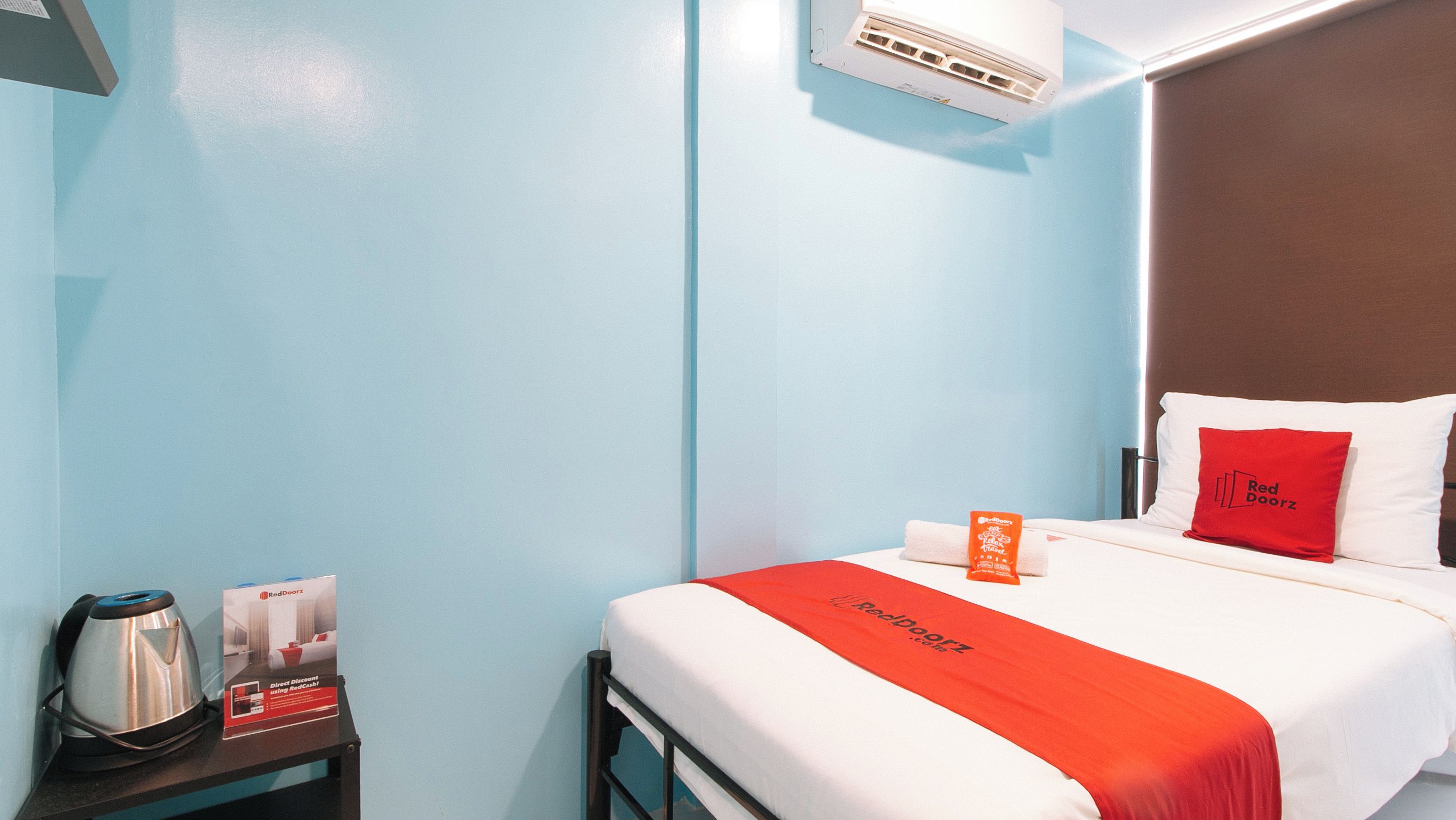 A hotel room with a bed, a pillow, a red blanket, a pillowcase, a bedside table, a kettle, and a RedDoorz sign.