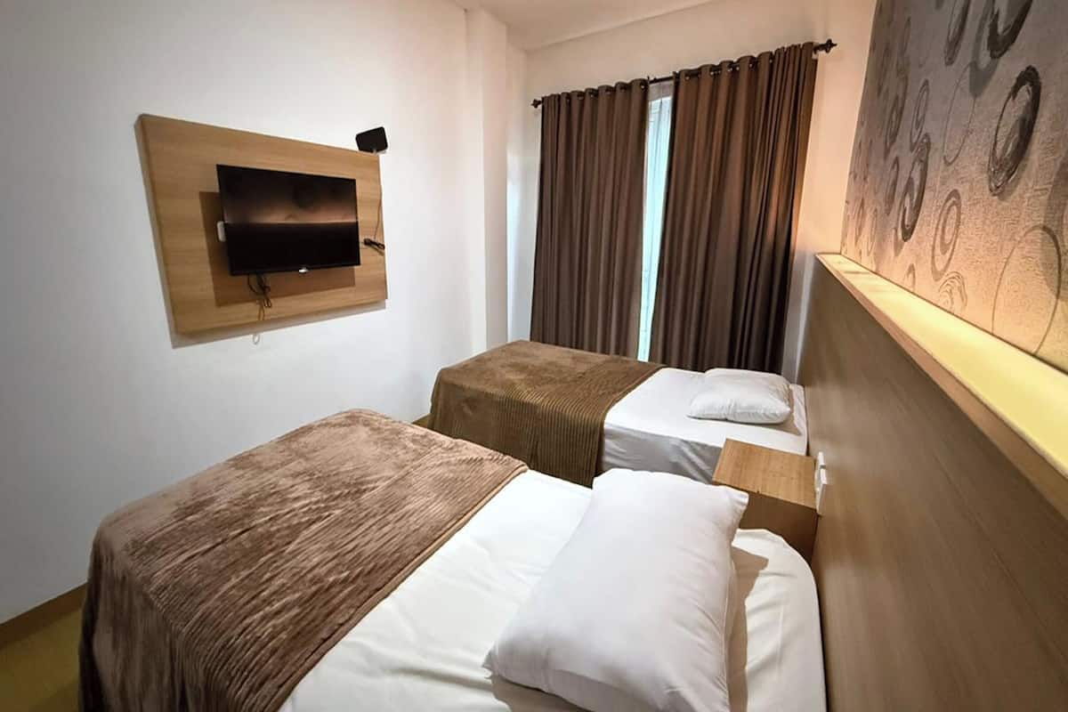 Ein Hotelzimmer mit zwei Einzelbetten, einem an der Wand befestigten Fernseher und einem großen Fenster mit Vorhängen.