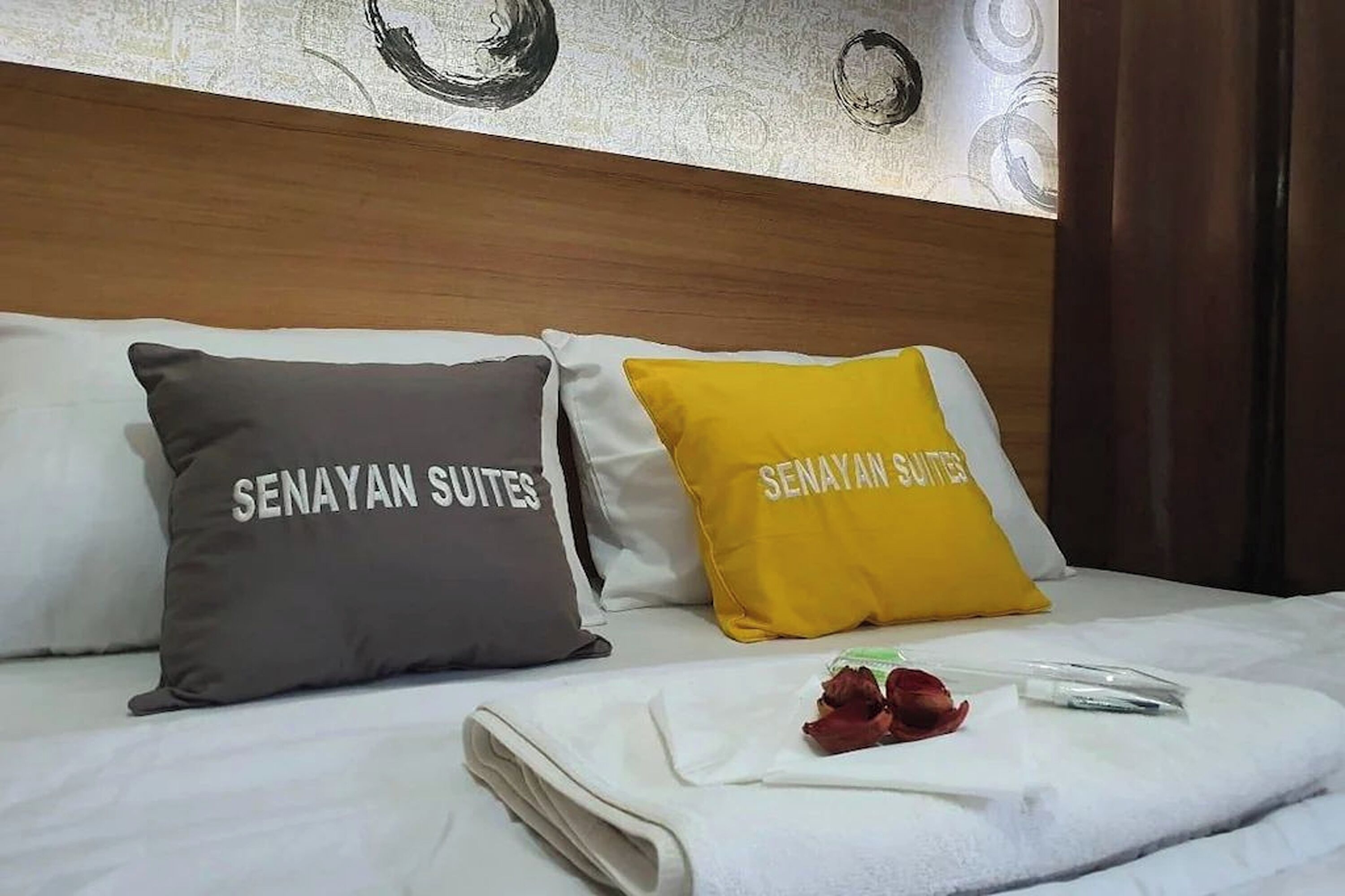 Un lit avec deux oreillers portant l’inscription « Senayan Suites » et une serviette blanche soigneusement pliée, ornée d’une rose rouge.