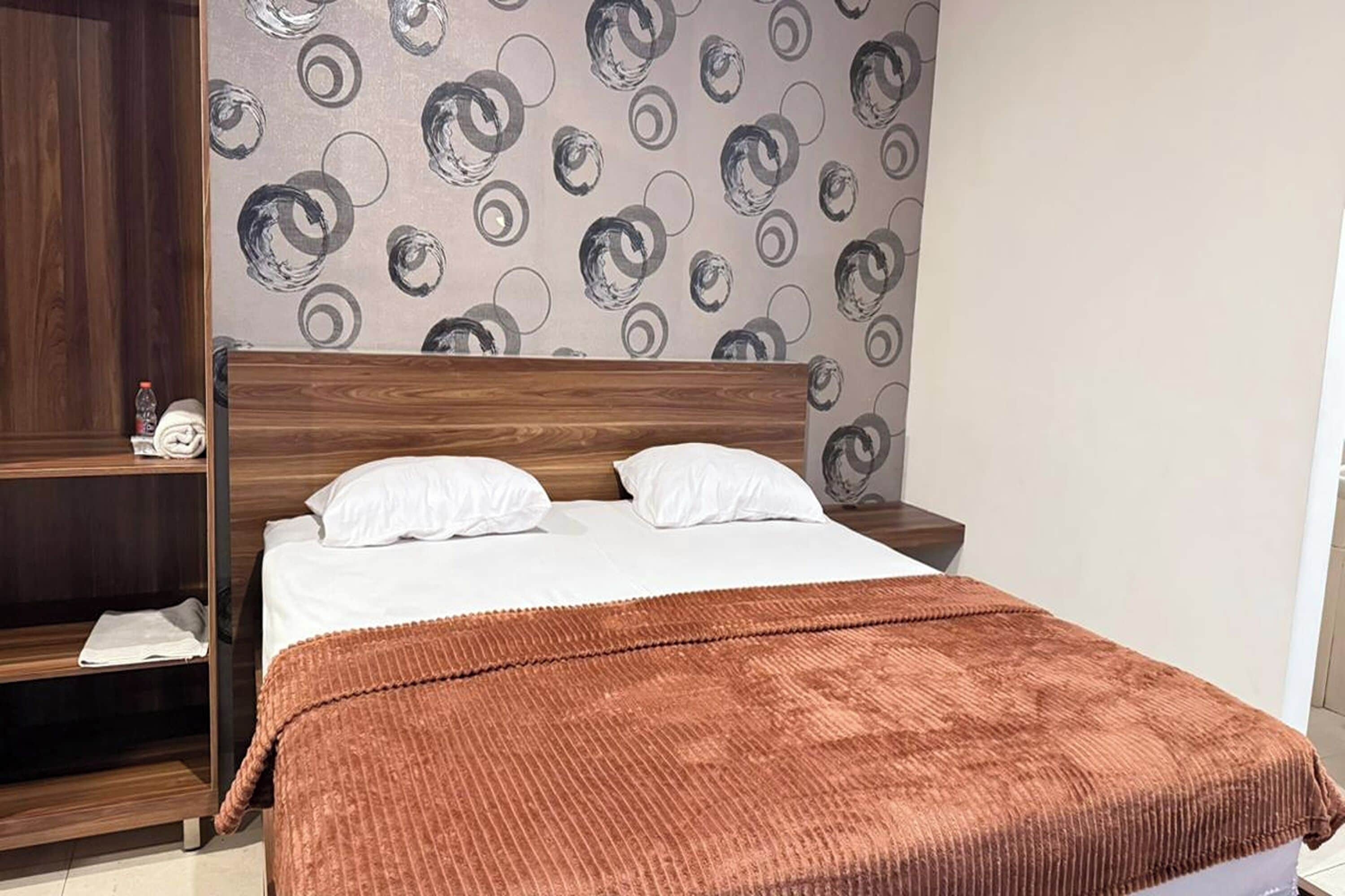 Un dormitorio con cama de madera, almohadas blancas, una manta marrón y una pared con diseño circular.