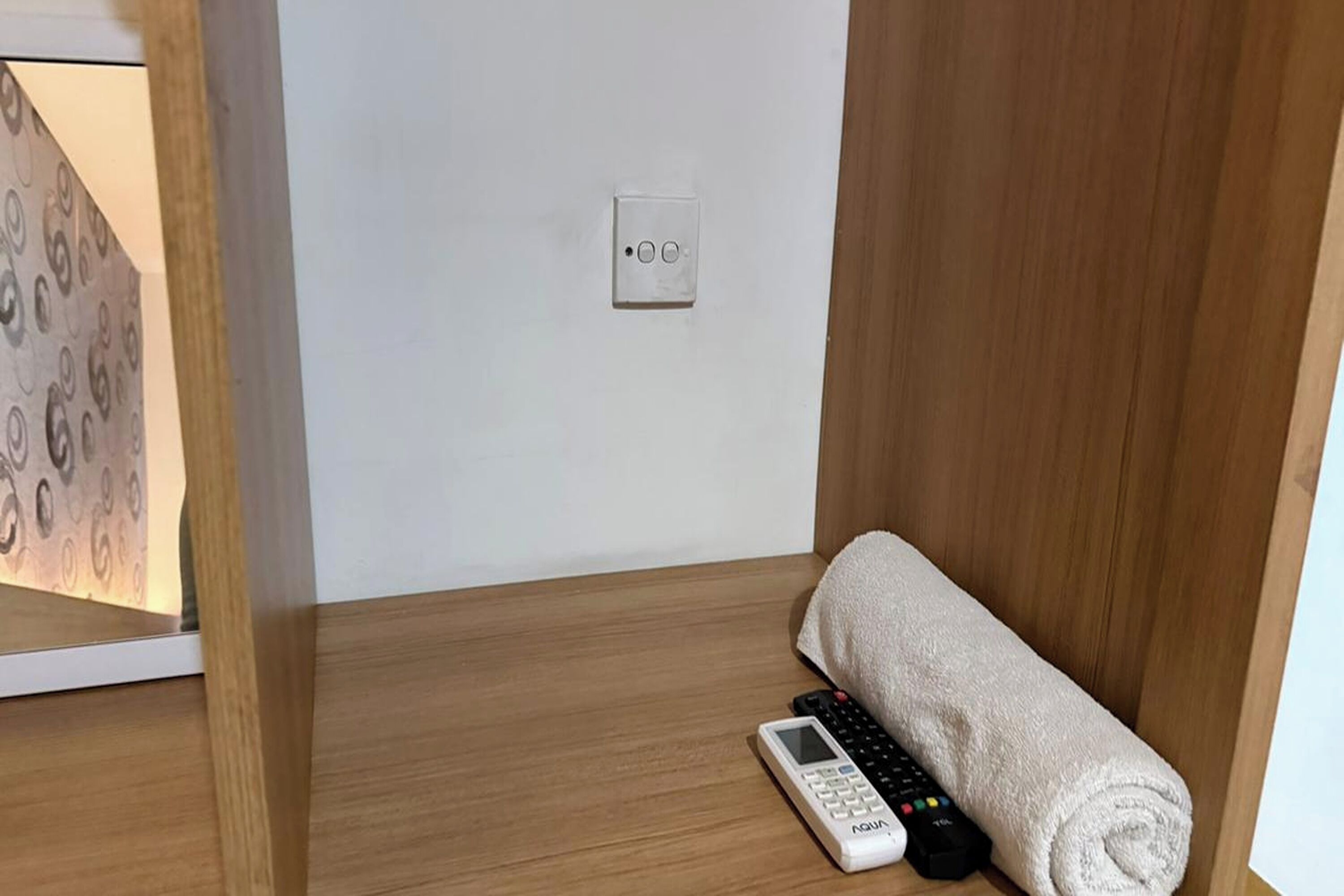 A hotel room with a wooden floor, a white wall, a power outlet, a remote control, and a rolled towel.
