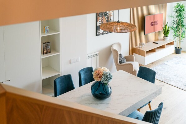 A modern living room with a dining area, featuring a marble table, blue chairs, a decorative vase, a hanging rattan light, and a TV on the wall.