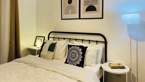 A bedroom with a bed, a bedside table with a lamp, a wall-mounted picture, and a nightstand with a framed photo.
