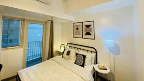 A bedroom with a bed, a bedside table, a wall-mounted lamp, and a window with a view of a building outside.