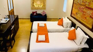A hotel room with two beds, a desk, a chair, a small table, and a wall-mounted lamp. - Hotel Dev Villa (Jaisalmer)