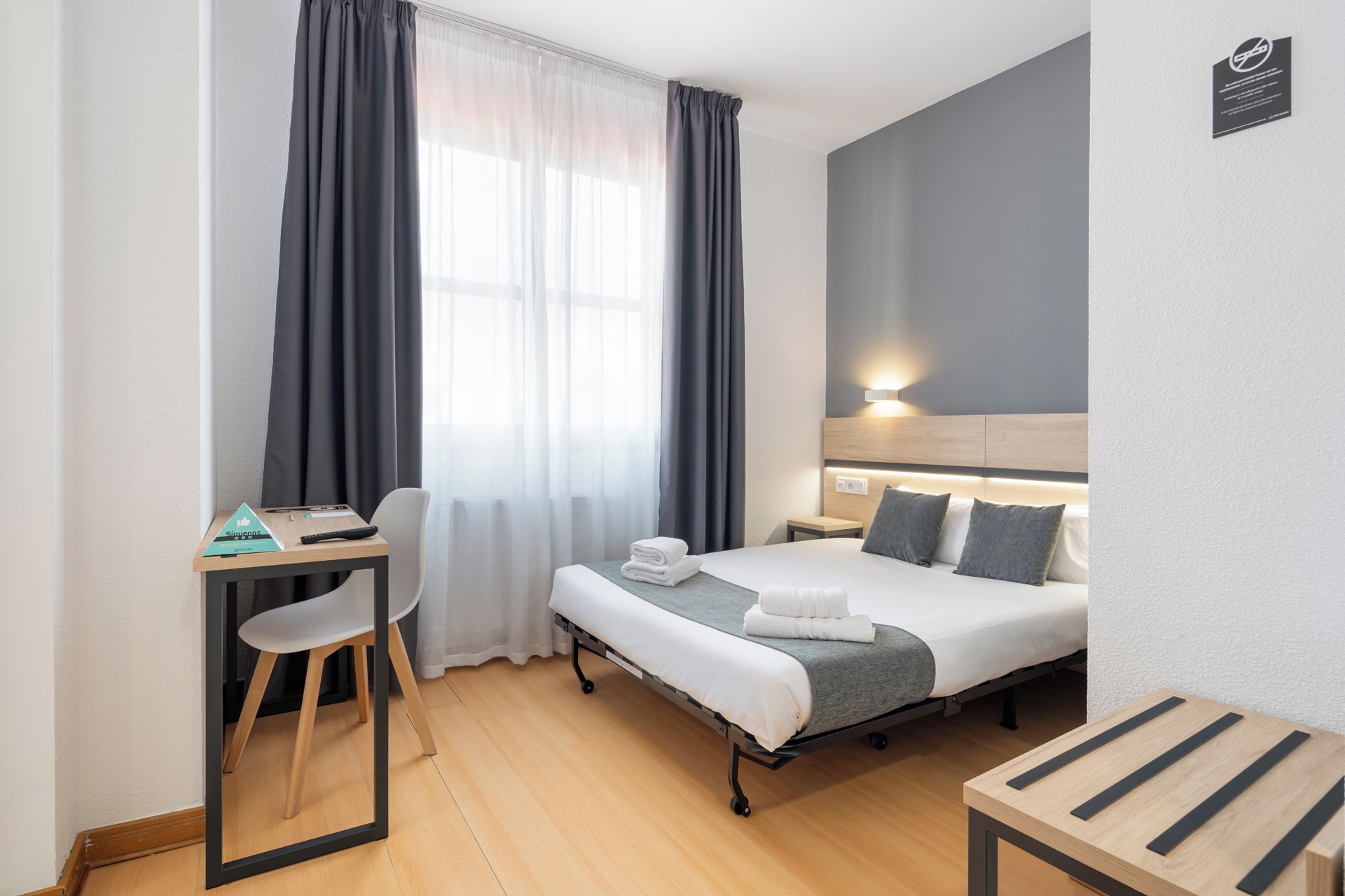 A hotel room with a bed, bedside table, chair, desk, and a window with curtains.