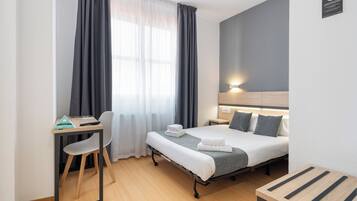 A hotel room with a bed, bedside table, chair, desk, and a window with curtains.