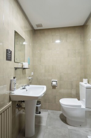 Ein Badezimmer mit weißem Waschbecken, Toilette und Spiegel. An der Wand befinden sich ein Mülleimer und eine Seifenspender.