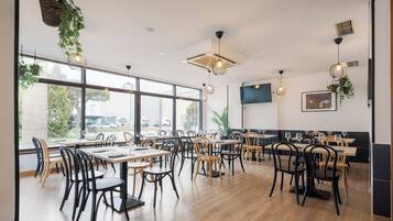 Un restaurante espacioso con mesas y sillas de madera, ventanas amplias, plantas colgantes e iluminación moderna.