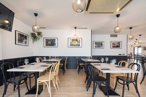 Ein modernes Restaurant mit langen Tischen, schwarzen Holzstühlen und Pendelleuchten.