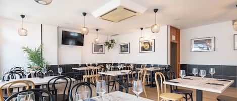 Interior de restaurante com várias mesas, cadeiras, luminárias pendentes e obras de arte nas paredes.