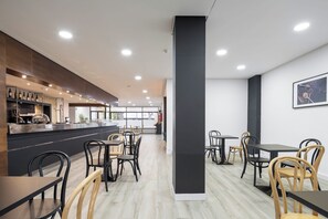 Egy modern kávézó, melynek központi eleme egy oszlop, a bútorzat pedig fából és feketéből áll, a pultján pedig borosüvegek sorakoznak.