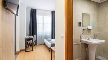 Una habitación de hotel con cama, escritorio, silla, televisión y baño con lavamanos y espejo.