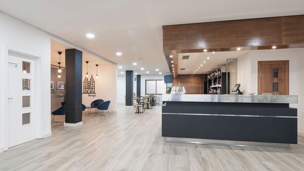 A modern hotel lobby with a central reception desk, seating area, and a view of the city. - Alda Puerto Seco (Burgos)