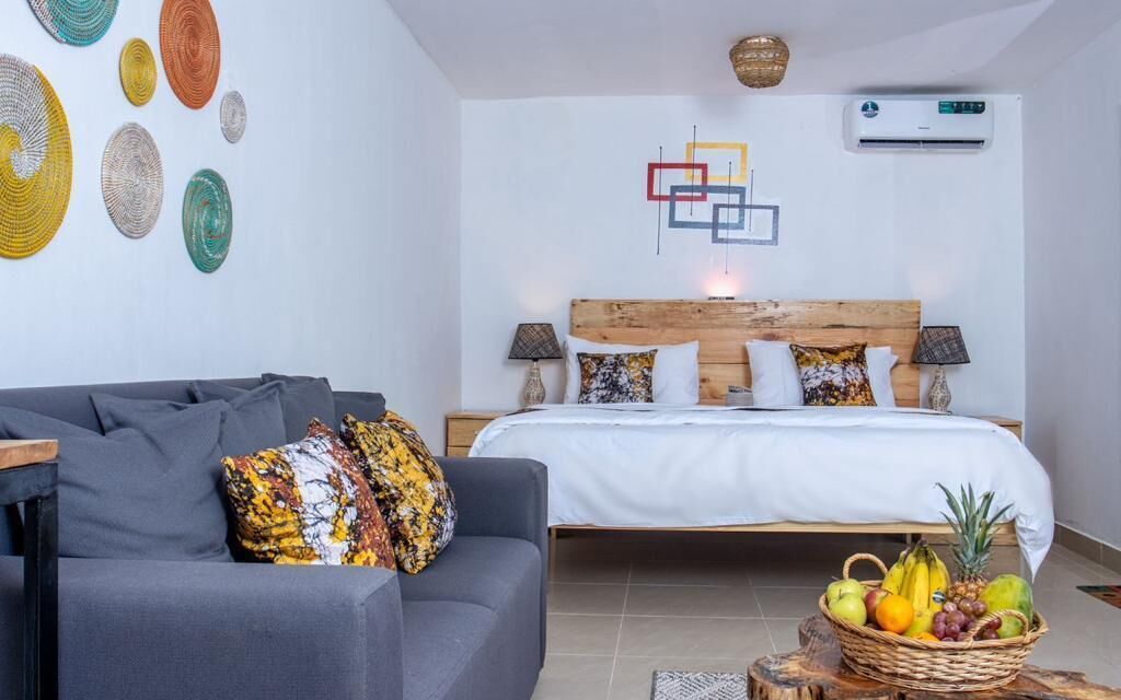 A bedroom with a bed, bedside tables, a gray sofa, a fruit basket, and wall art.
