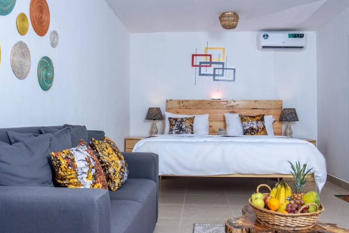 Une chambre à coucher avec un lit, des tables de chevet, un canapé gris, un panier de fruits et des œuvres d’art murales.