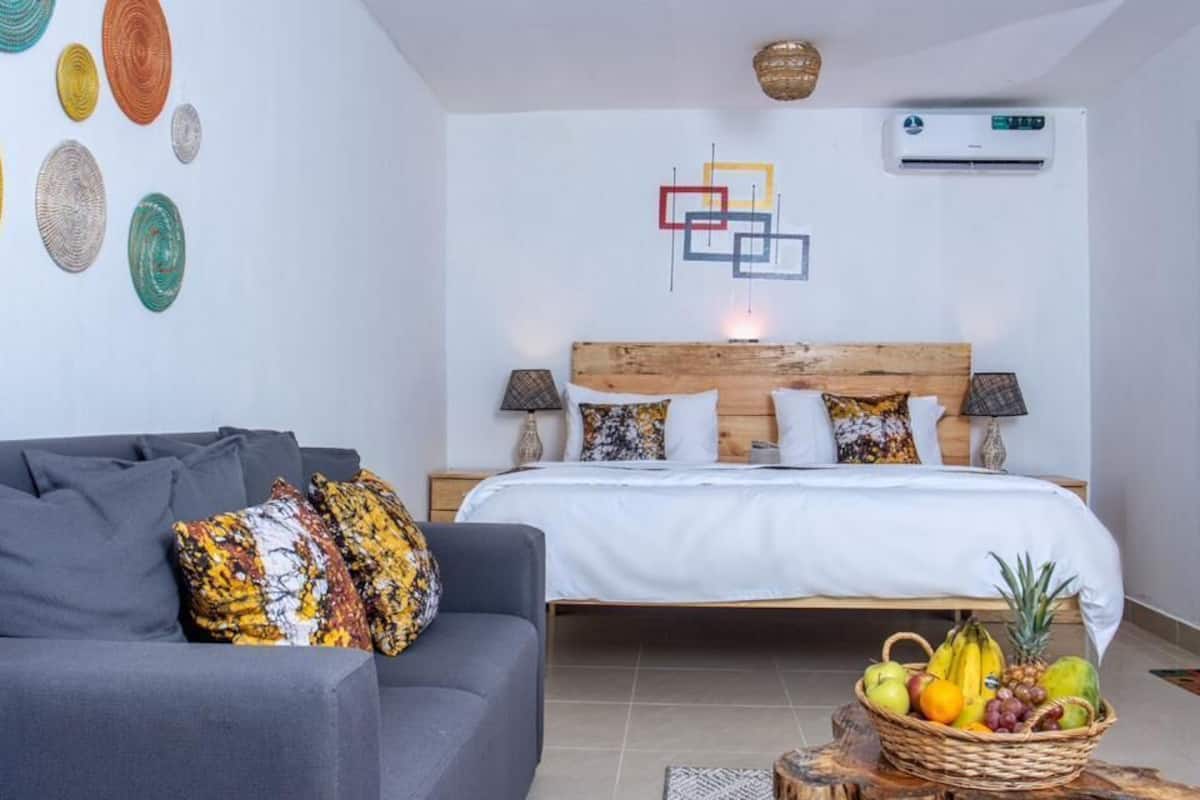 A bedroom with a bed, bedside tables, a gray sofa, a fruit basket, and wall art.