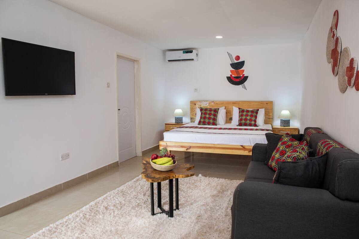 Une chambre à coucher avec un lit, une télévision, une table de chevet avec une lampe, une petite table ronde avec des fruits et un canapé.