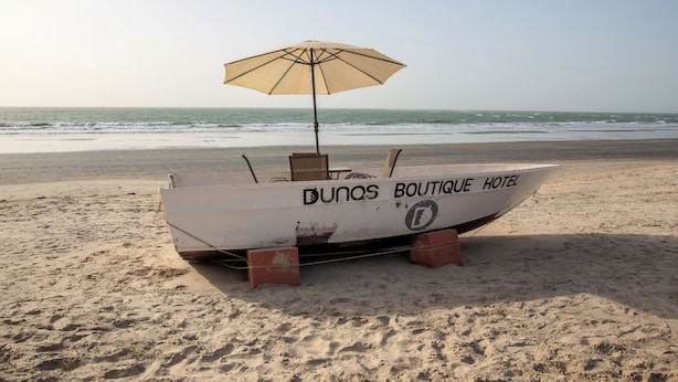 A boat with "DUNOS BOUTIQUE HOTEL" written on it, parked on a sandy beach with the ocean in the background.
