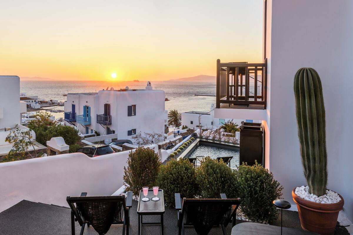 Eine Terrasse mit Meerblick bei Sonnenuntergang, weiße Gebäude, ein Kaktus und Sitzgelegenheiten im Freien.