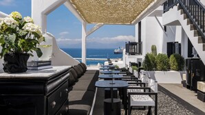 Een terras met tafels en stoelen, een wit gebouw en uitzicht op de oceaan.