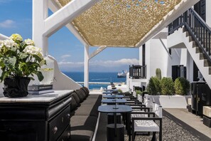 An outdoor seating area with tables and chairs, a white building, and a view of the ocean. - She Mykonos Boutique Hotel (Mykonos)