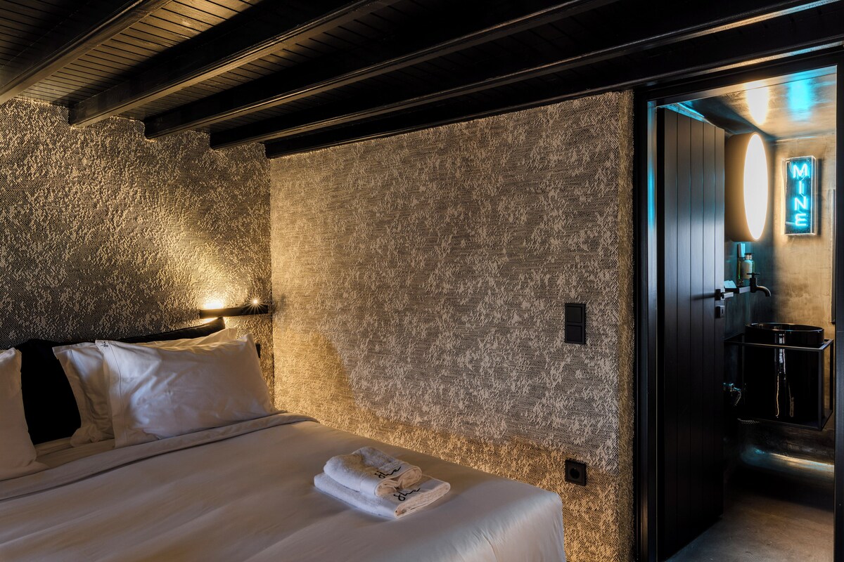 Een hotelkamer met een structuurwand, een net opgemaakt bed met witte lakens, en een zichtbare badkamer met een wastafel en spiegel.