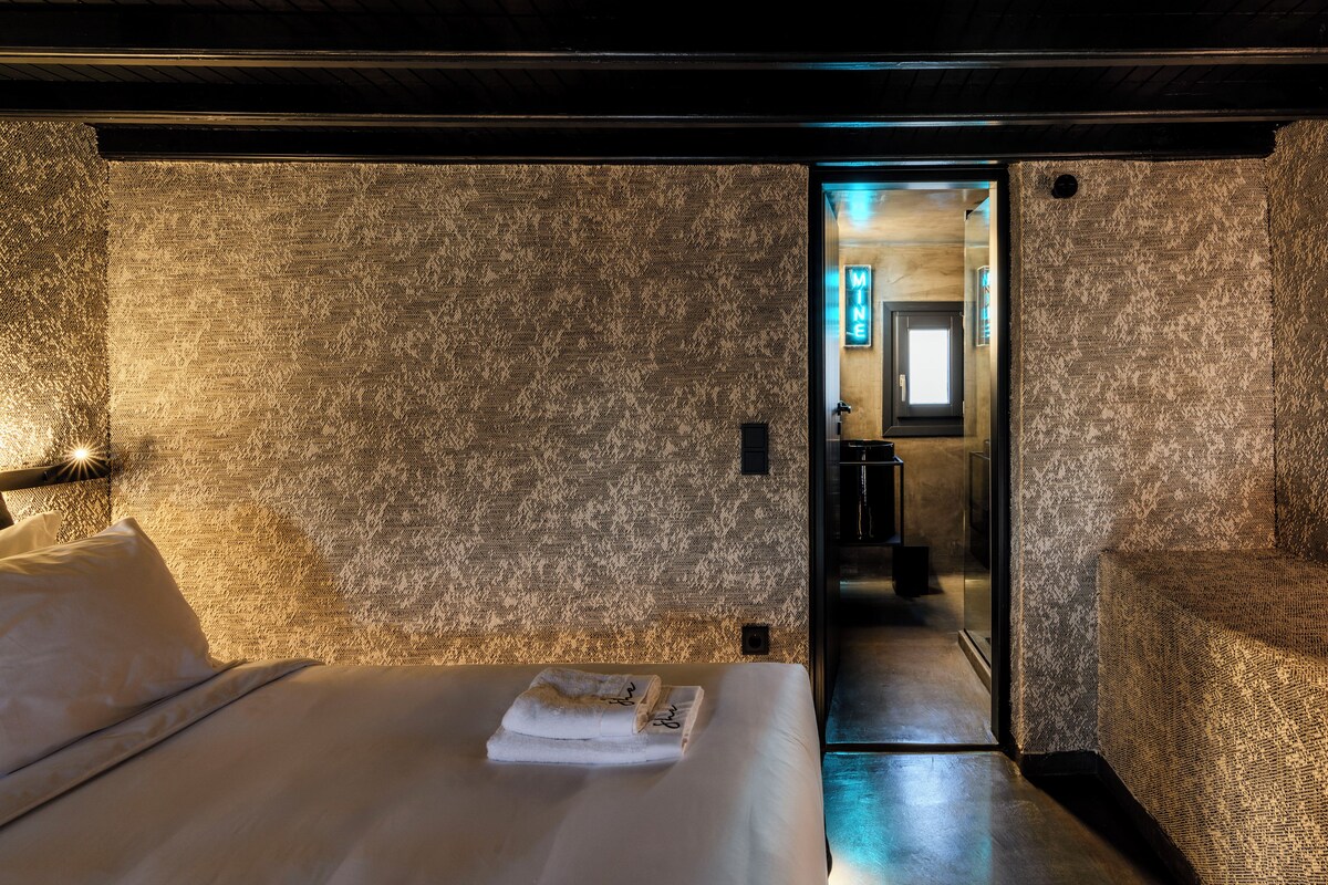 Ett hotellrum med en vägg som har en intressant yta, en snyggt bäddad säng med en handduk på, och en dörröppning som leder till ett badrum.
