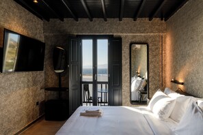 Quarto com cama, televisão, espelho grande e vista para o mar através de portas abertas.