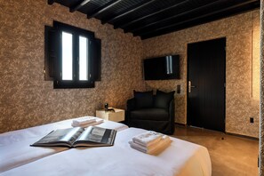 Une chambre d’hôtel avec un grand lit, une fenêtre avec des volets, un téléviseur à écran plat fixé au mur, un fauteuil noir et une table d’appoint en bois avec une lampe.