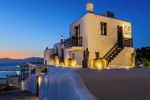 A modern white building with a balcony and staircase, located near a body of water with a clear sky. - She Mykonos Boutique Hotel (Mykonos)