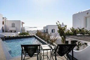 A rooftop pool with two chairs and a table, overlooking a cityscape and the sea.