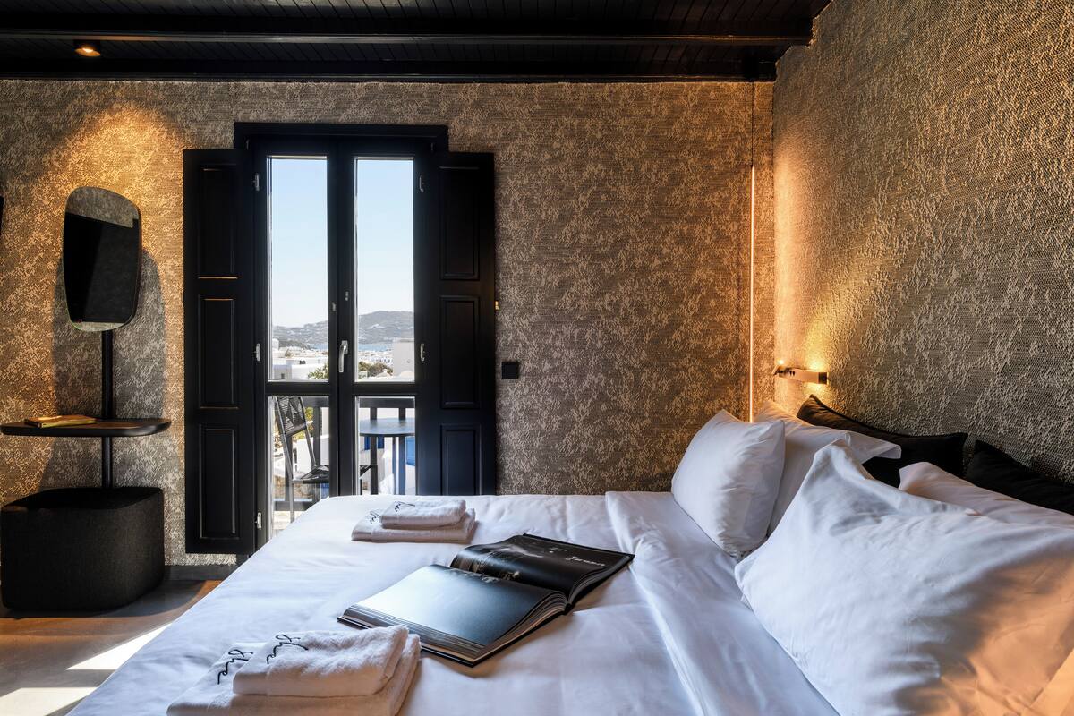 Ein Hotelzimmer mit einem Bett, einem Buch, einem Handtuch, einem schwarzen Stuhl und einem an der Wand befestigten Fernseher.