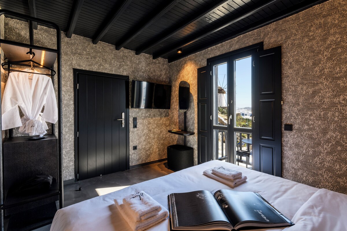 Une chambre d’hôtel moderne avec un grand lit, un téléviseur à écran plat fixé au mur et une vue sur la ville, visible à travers la porte ouverte.