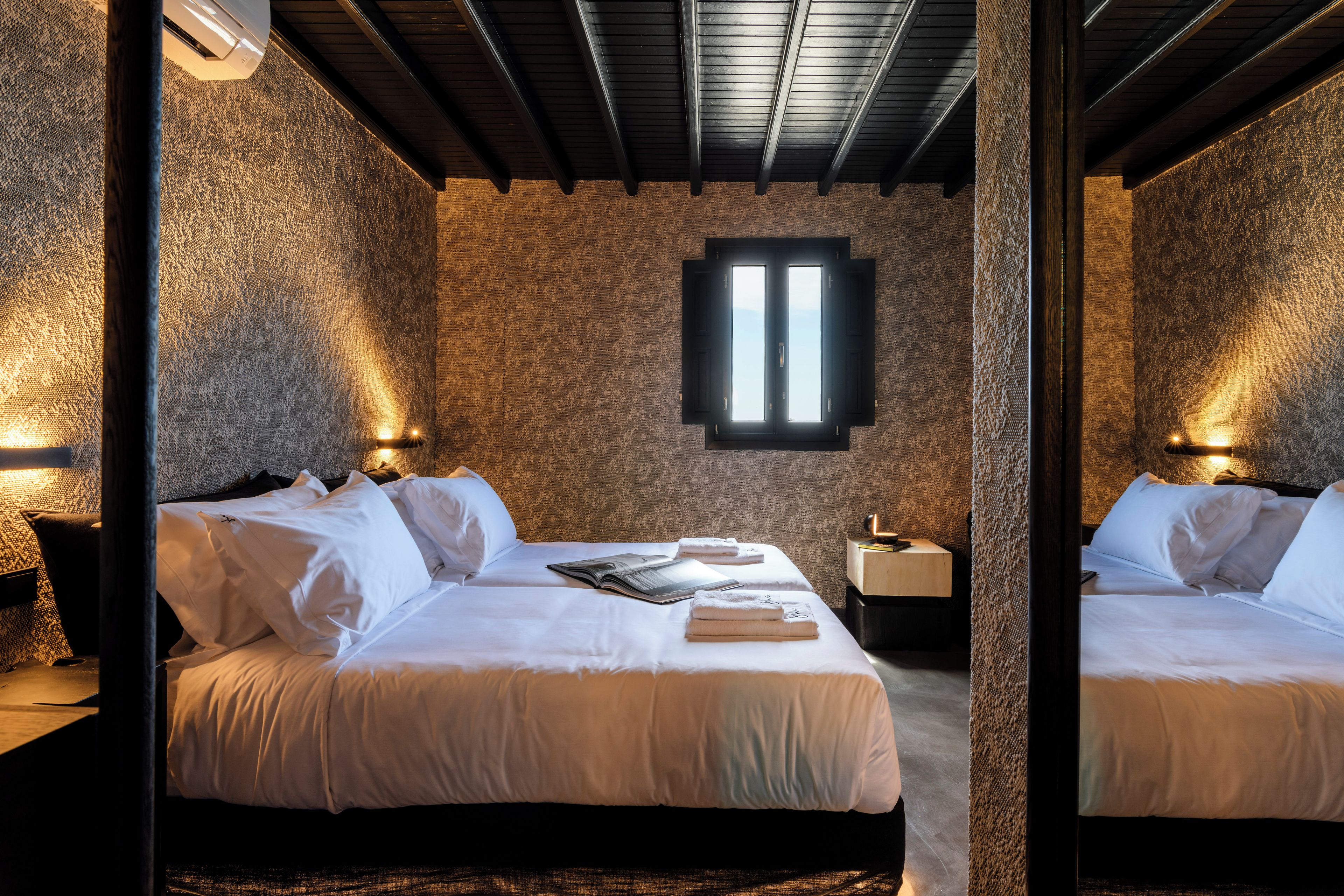 A room with two beds, a stone-textured wall, a window with blinds, and wall-mounted lights.