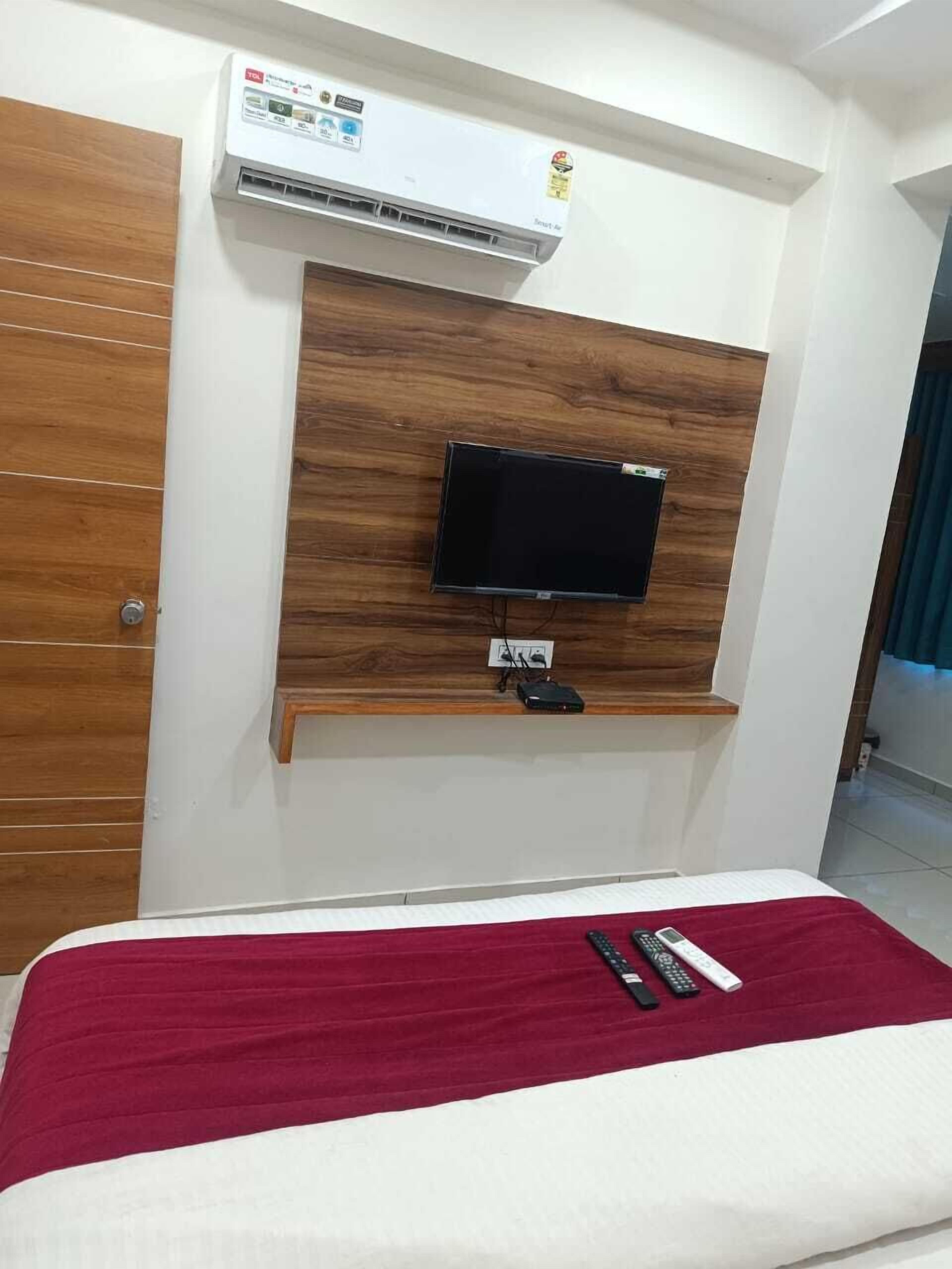 Quarto de hotel com cabeceira de madeira, televisão montada na parede, cama com manta vermelha e controle remoto sobre a cama.